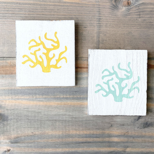 Two square white wooden signs with coral designs, one in yellow and one in mint, displayed on a rustic wooden surface.