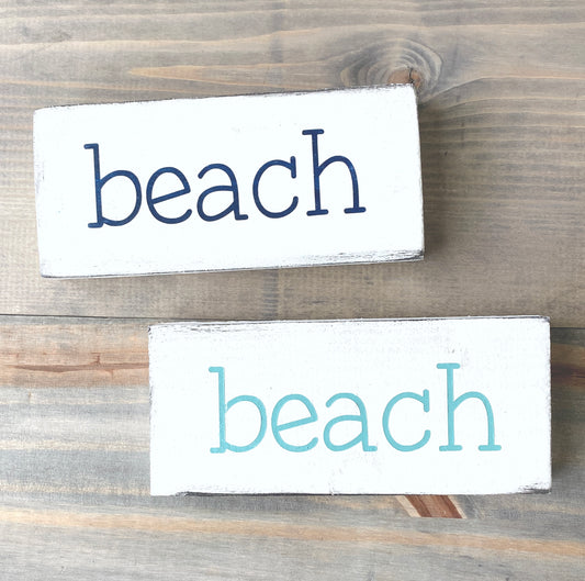 Two rectangular white wooden signs with the word "beach" printed in lowercase letters, one in dark navy and one in aqua, displayed on a rustic gray-brown wooden surface.