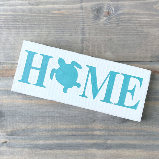 A rectangular white sign with the word “HOME” in aqua, where the letter O is replaced by an aqua sea turtle silhouette, placed on a rustic gray-brown wooden surface.
