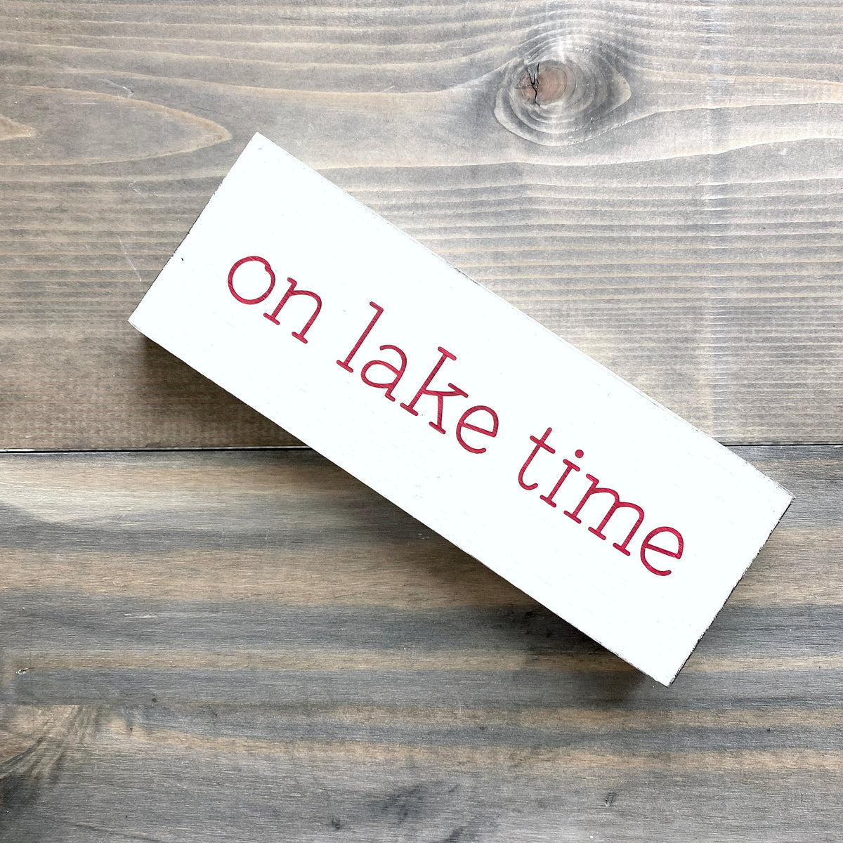 A white rectangular wooden sign with the phrase 'on lake time' in red lowercase lettering, photographed at an angle on a rustic gray-brown wood plank surface.