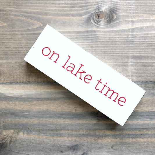 A white rectangular wooden sign with the phrase 'on lake time' in red lowercase lettering, photographed at an angle on a rustic gray-brown wood plank surface.
