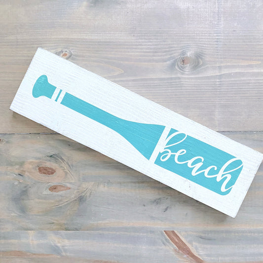 Wooden sign with a blue oar design and 'beach' text on a wooden surface