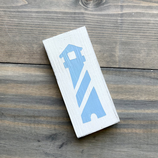 A rectangular white wooden block painted with a light blue lighthouse design, resting on a rustic dark wood surface.