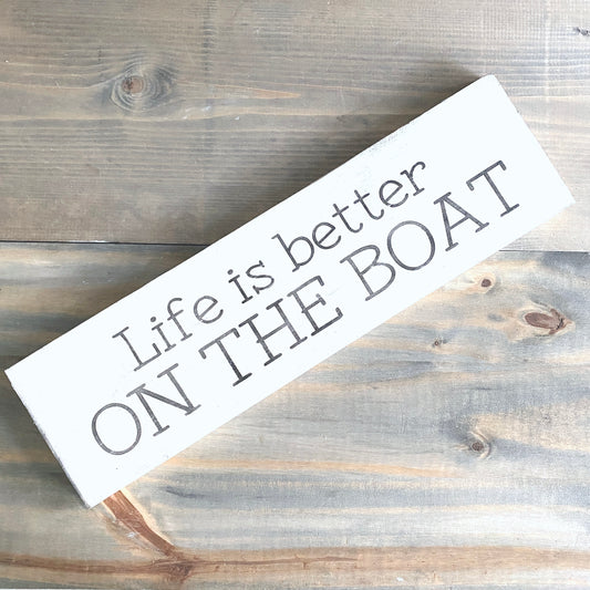 A white rectangular wooden sign with the phrase 'Life is better ON THE BOAT' in black lettering, photographed diagonally on a rustic gray-brown wood plank surface.