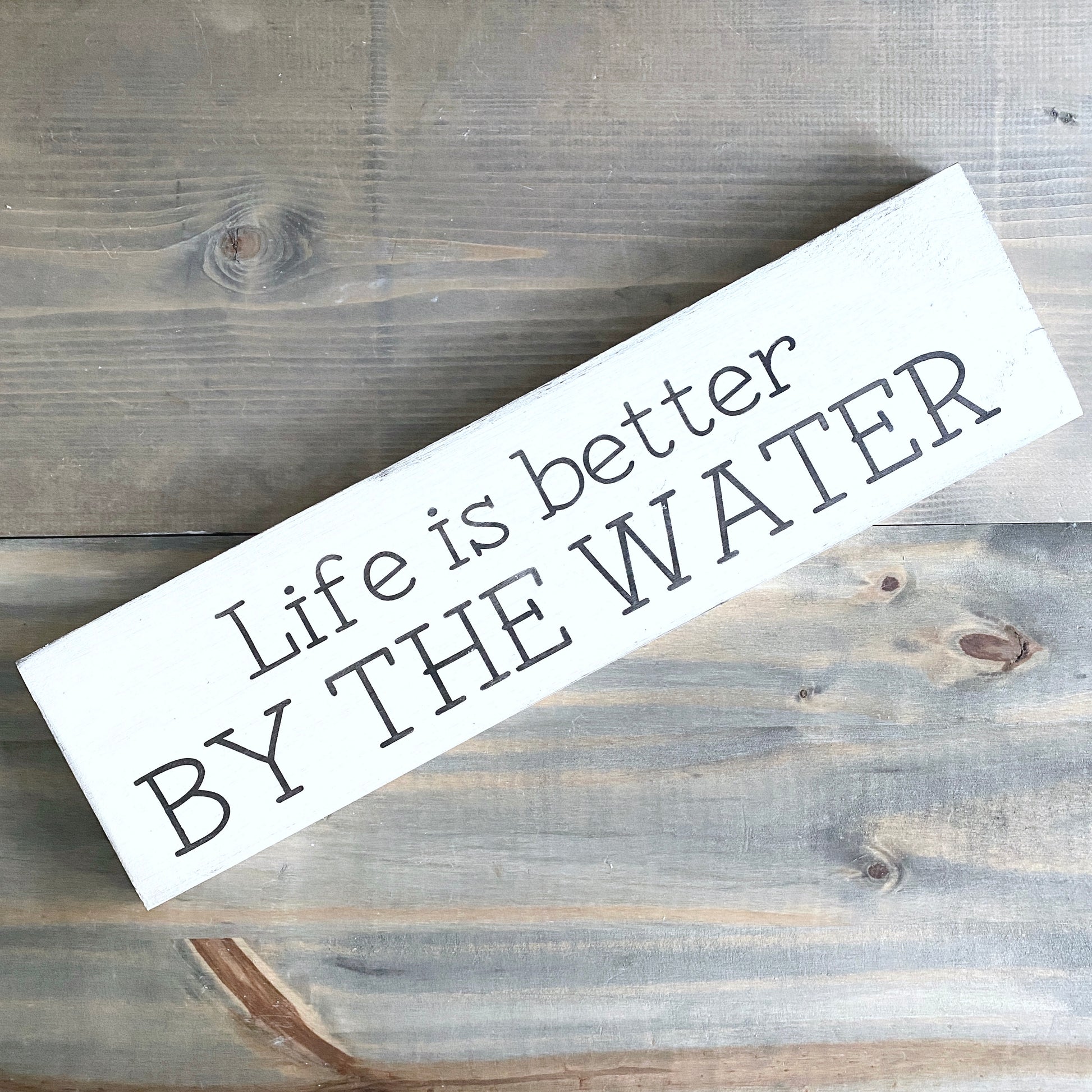 A rectangular white wooden sign with the phrase 'Life is better BY THE WATER' in dark gray lettering, photographed diagonally on a rustic gray-brown wood plank surface.