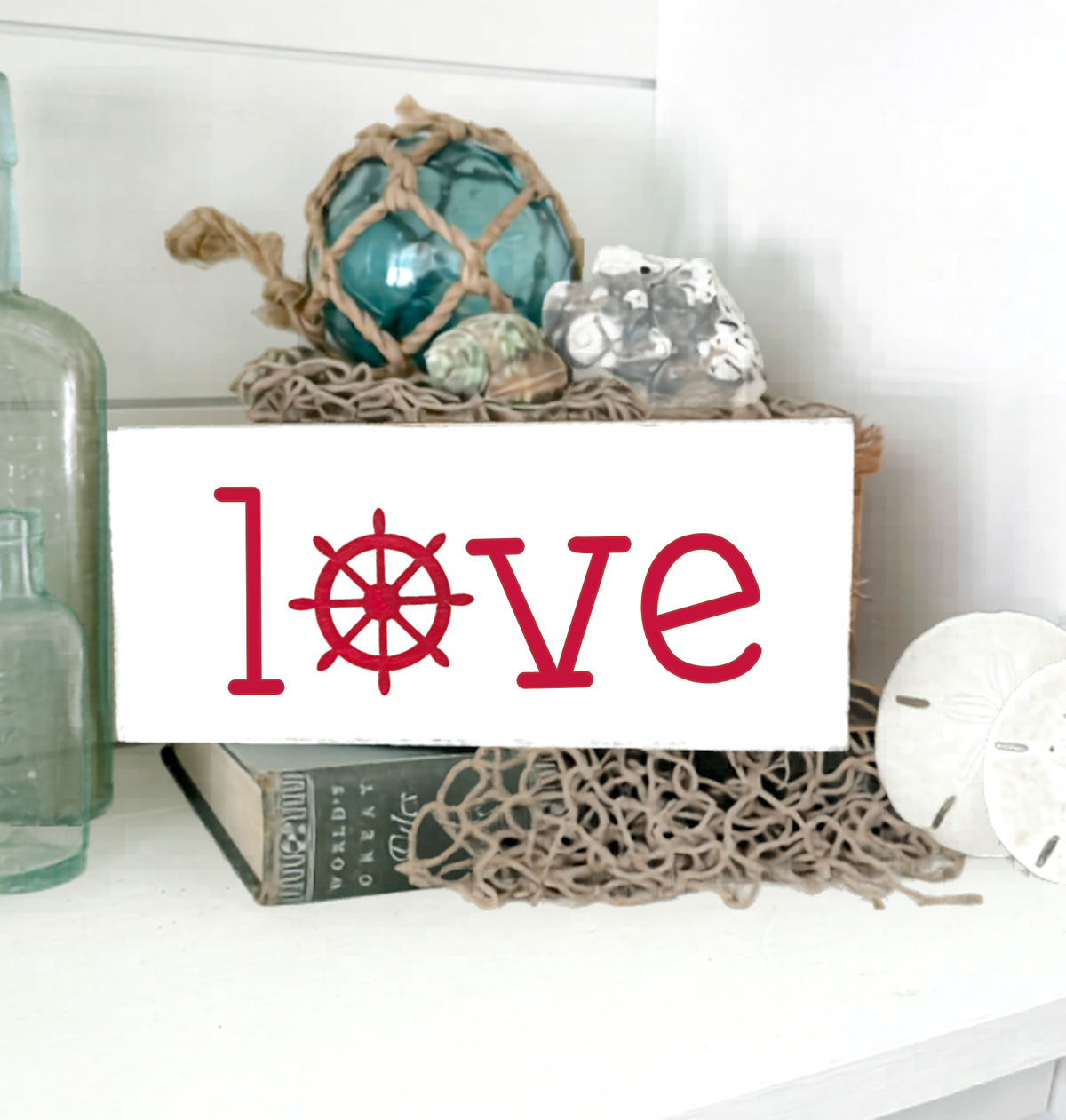 A white rectangular decorative sign with the word "love" in red, where the "o" is replaced by a red ship's wheel, displayed on a shelf with nautical decor including a blue glass orb, rope, crystals, a book, and sand dollars.