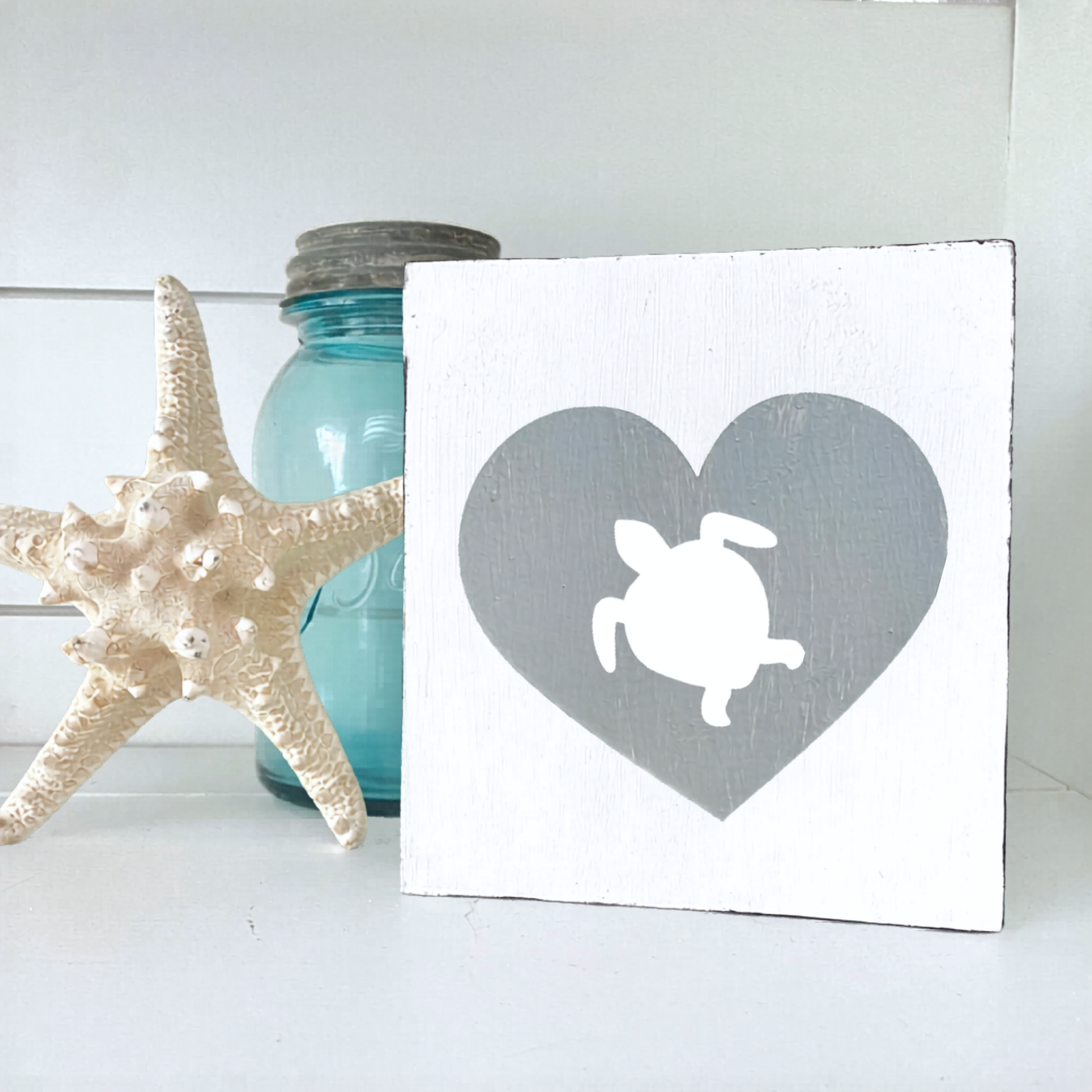 A square white wooden sign with a gray heart and white turtle silhouette, styled on a white surface next to a starfish and a blue glass jar.