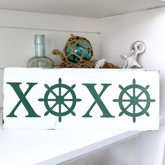 A white rectangular sign, featuring the letters X O X O in dark green with ship's wheel motifs. The sign is displayed on a white shelf alongside nautical-themed decor, including a glass bottle, fishing float, and a white anchor.
