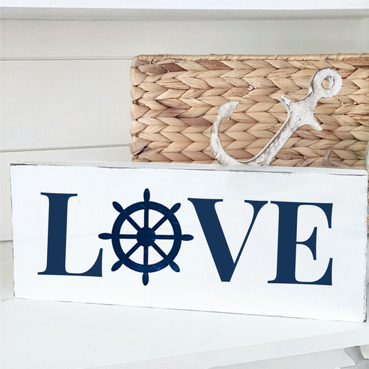 A white rectangular decorative sign with the word LOVE in navy blue, where the O is replaced by a navy ship's wheel. A white anchor decoration and a woven basket are placed behind it on a white shelf.