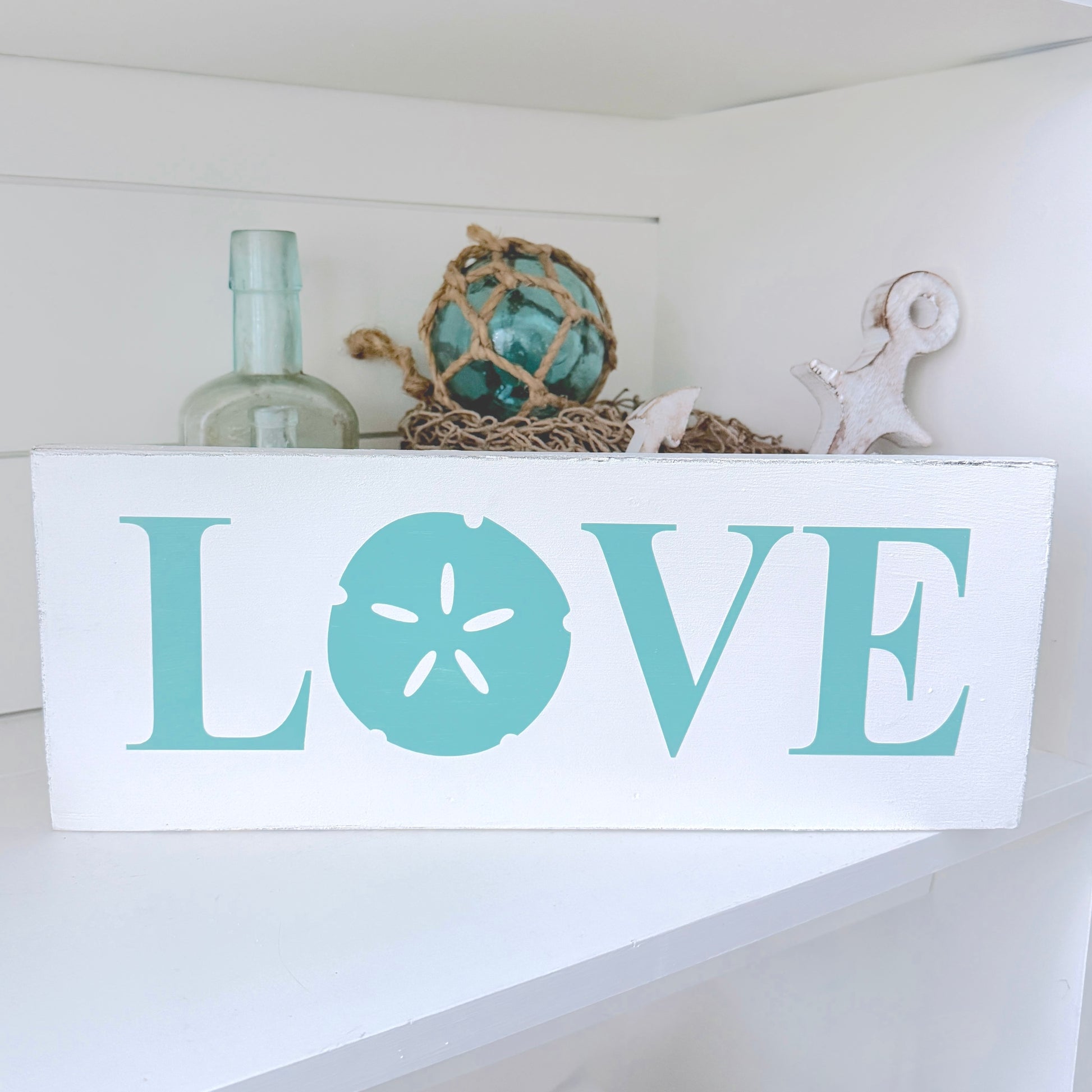 A white rectangular wooden sign with the word LOVE in large teal letters, where the O is shaped like a sand dollar. The sign sits on a white shelf with nautical decor behind it, including a glass bottle, rope ball, starfish, and shell.