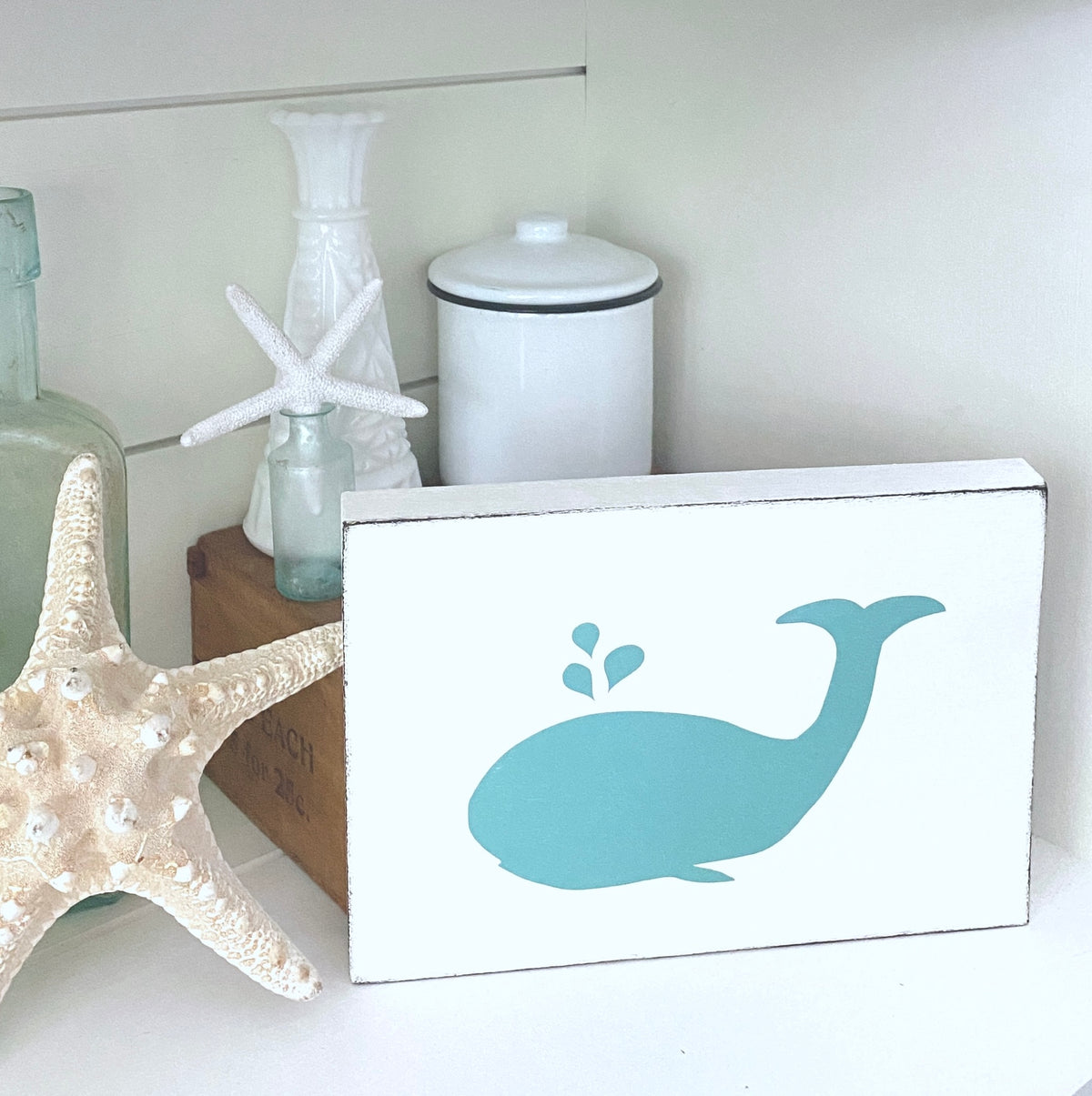 A square white decorative sign with a teal whale silhouette, styled on a shelf beside a starfish, glass bottles, and a white canister.