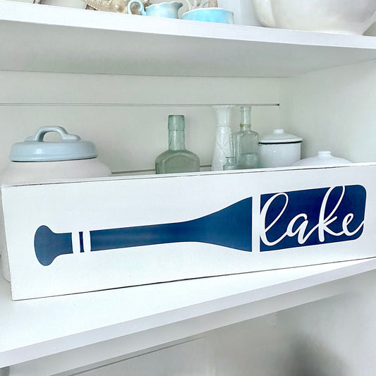 A white sign with a navy blue oar silhouette and the word "lake" in white script, displayed on a white shelf with glass bottles and ceramic containers behind it.