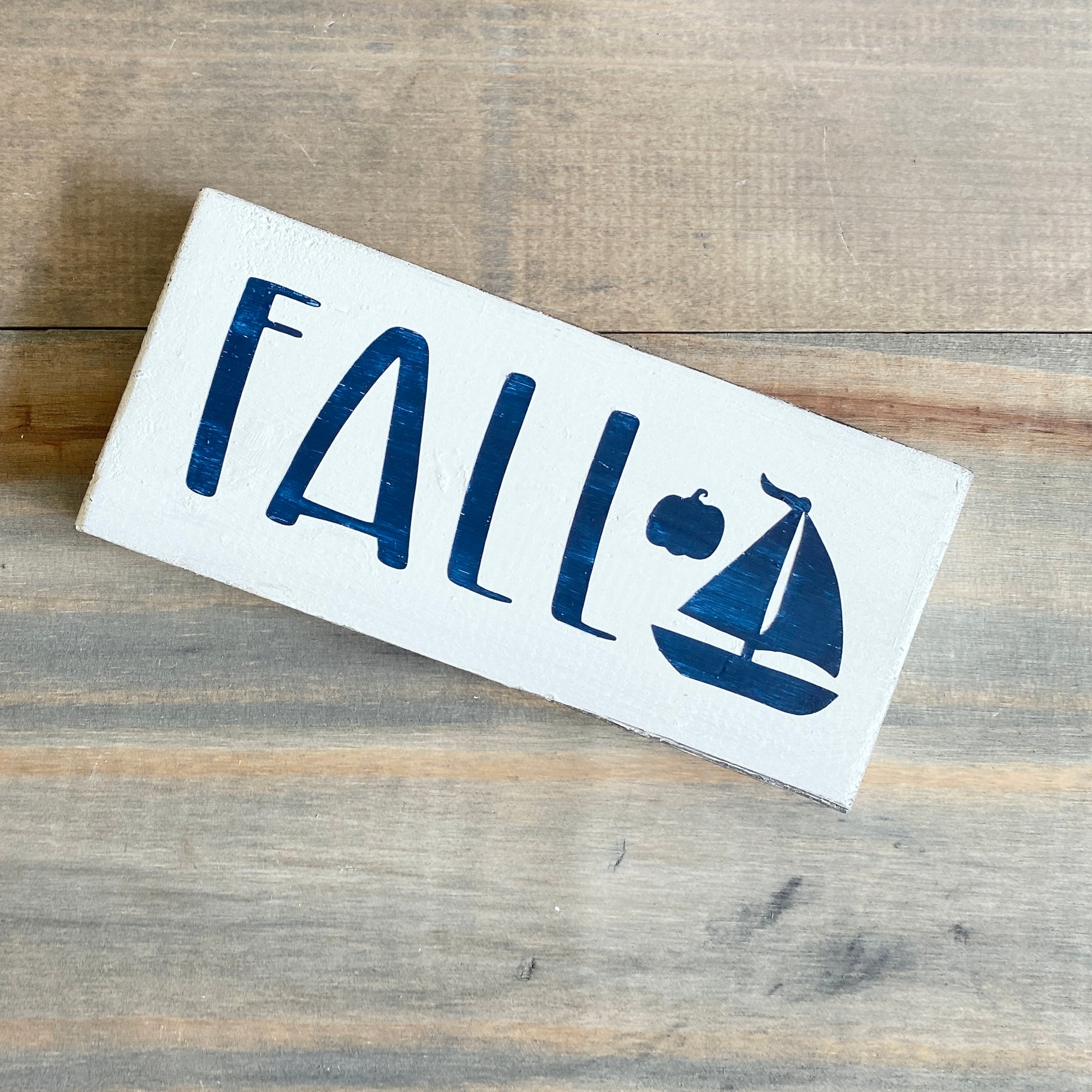 Rectangular sign with 'FALL' and a sailboat on a wooden surface
