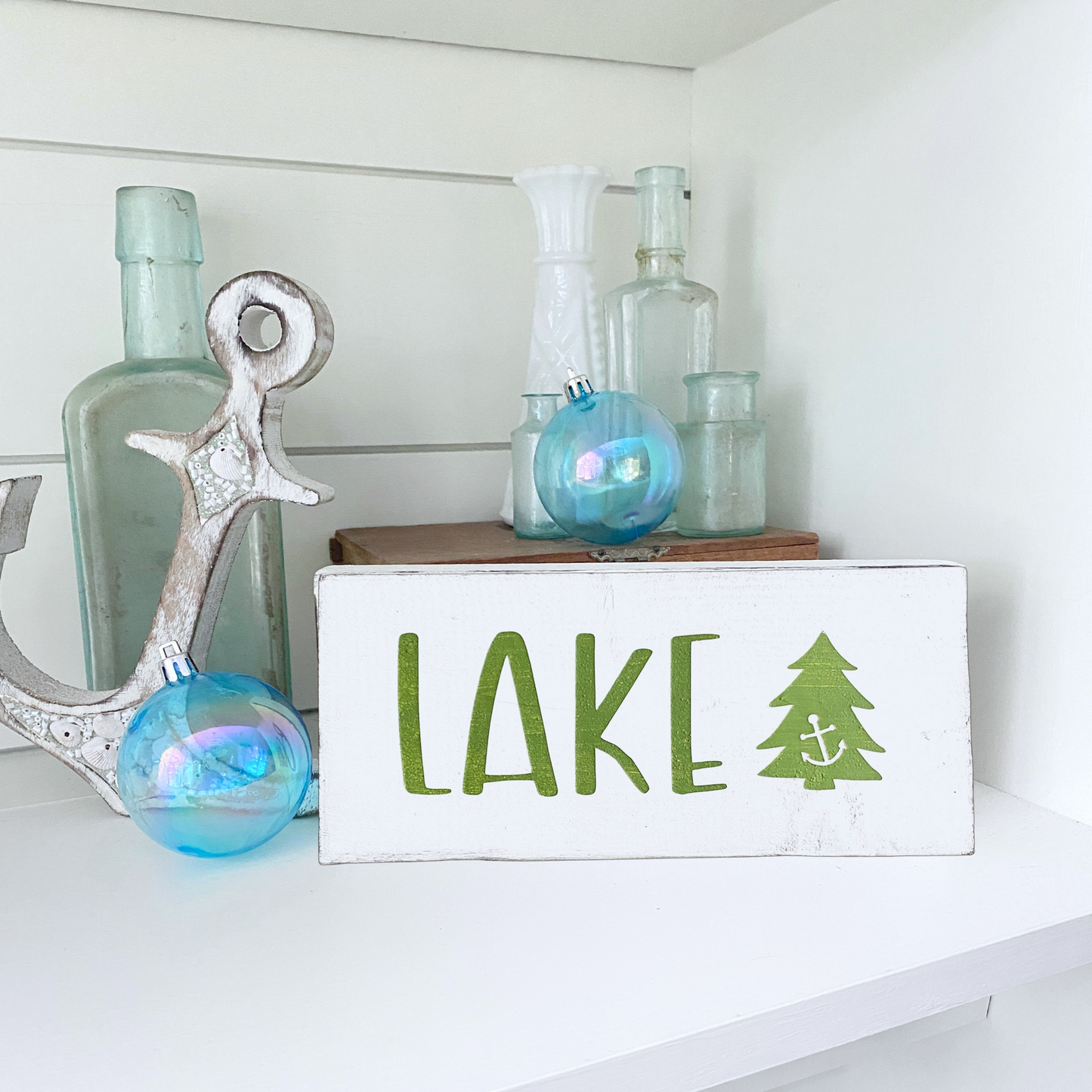 A white rectangular decorative sign with the word "LAKE" in green and a green tree with a small anchor inside, styled on a white shelf with glass bottles and blue ornaments.