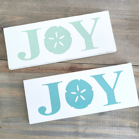 Two white rectangular decorative signs with the word "JOY" in large pastel letters, one in mint green and one in  aqua blue, displayed on a rustic wooden surface.