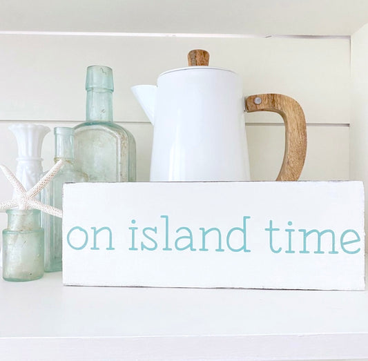 A white rectangular wooden sign with the words 'on island time' in light blue lowercase letters, styled on a white shelf with a white ceramic pitcher, glass bottles, and a starfish.