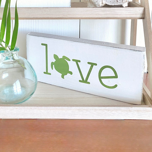 Decorative sign with 'love' and sea turtle design on a wooden shelf displayed beside a vase and palm frond