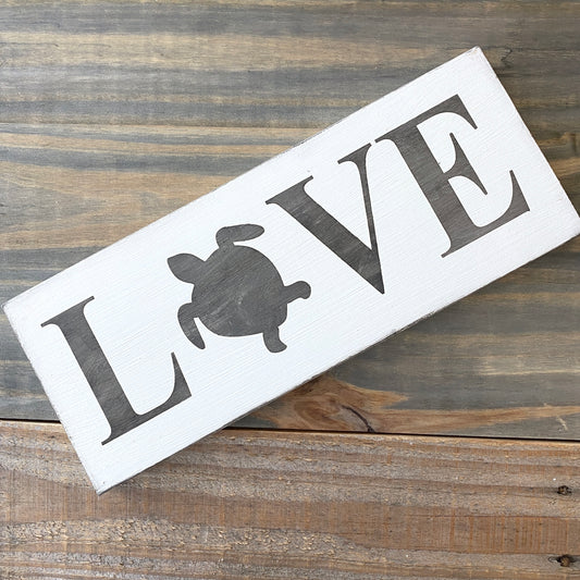 A rectangular white wooden sign with the word LOVE, where the O is replaced by a dark gray sea turtle silhouette, displayed diagonally on a rustic wooden surface.