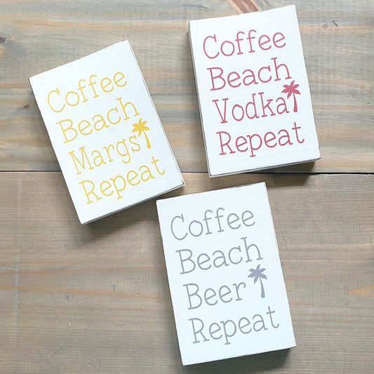 Three signs with humorous text designs on a wooden surface, coffee beach repeat signs