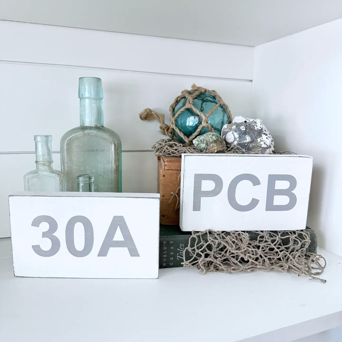 White wooden shelf signs with gray lettering, one reading '30A' and the other 'PCB', styled on a white shelf with glass bottles, nautical netting, shells and a wooden box.