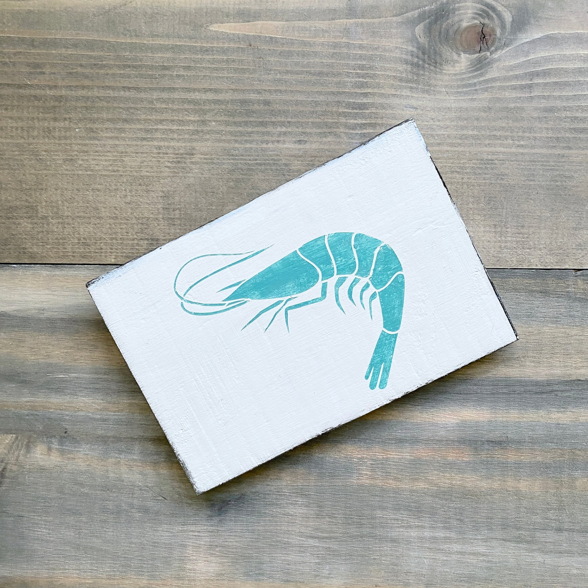 A rectangular white sign featuring an aqua painted shrimp illustration, photographed on a rustic gray-brown wooden surface.