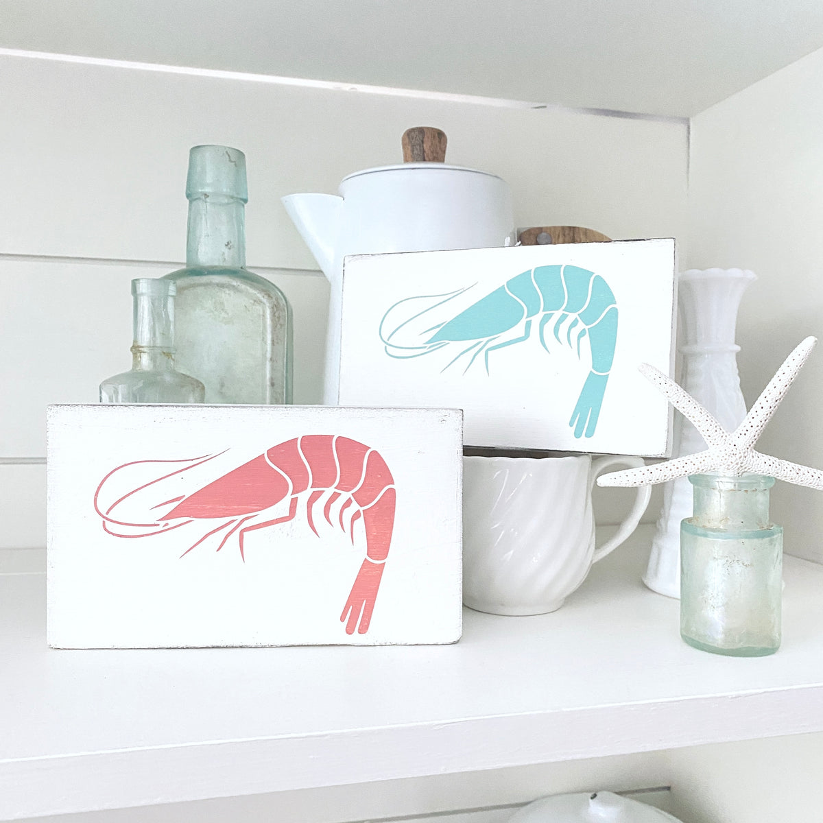 Two white rectangular decorative signs with painted shrimp designs in aqua and pink, displayed on a white shelf among glass bottles, a white teapot, a mug, and a starfish.