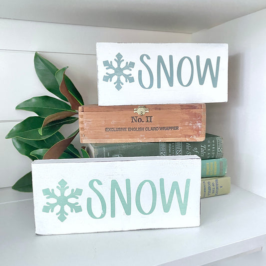 Two white wooden decorative signs with the word 'SNOW' in mint green and a snowflake icon, stacked on top of books with a green plant in a white shelf setting.