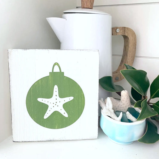 A square white wooden sign with a distressed finish, featuring a green circular ornament design with a white starfish in the center. The sign is styled on a white surface next to a white teapot with a wooden handle, a small light blue bowl with white coral, and green leaves.