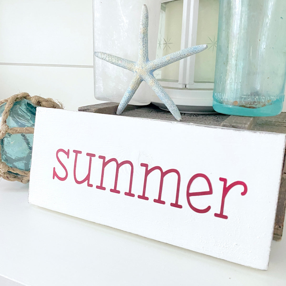 A white rectangular wooden sign with the word 'summer' in red lowercase script, styled with a starfish, glass jar, and rope decor on a white surface.