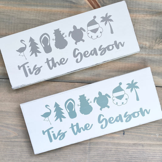 Two decorative signs with 'Tis the Season' text and holiday-themed icons on a wooden surface.