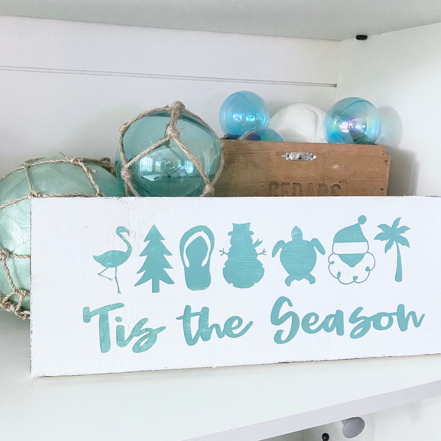 Decorative sign with icons and 'Tis the Season' text on a white shelf with decorative items.